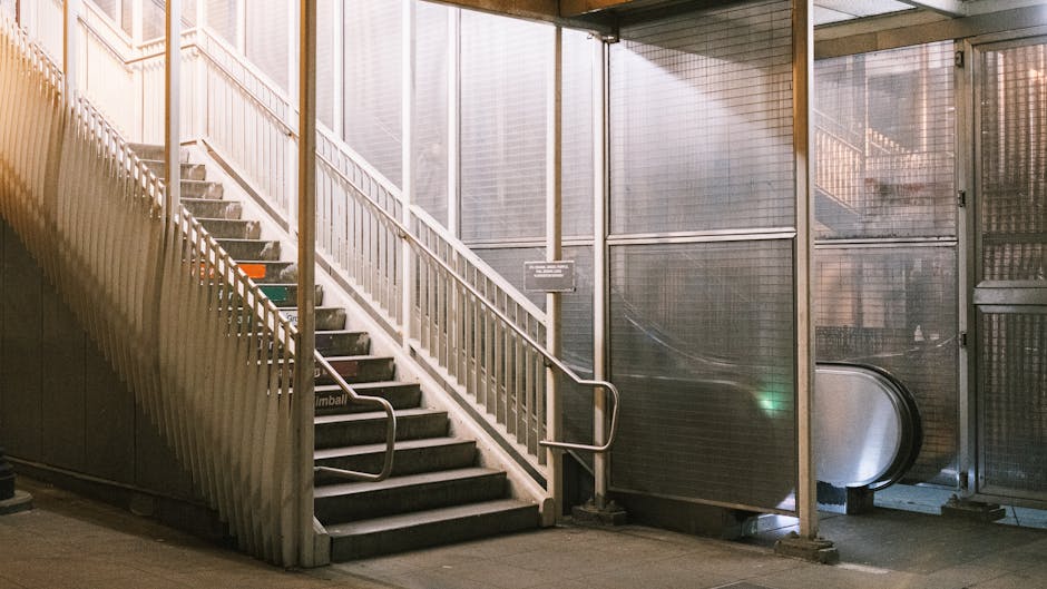A wide set of marble stairs leading upwards inside a building, with glass-paneled railings on both sides. The stairwell features a wooden ceiling with visible horizontal beams and a green directional 