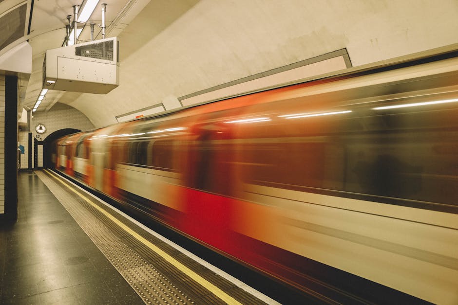 A moving train with a red and white exterior is captured in motion as it speeds through an underground station, creating a blurred effect. The station has a curved ceiling with beige panels and visibl